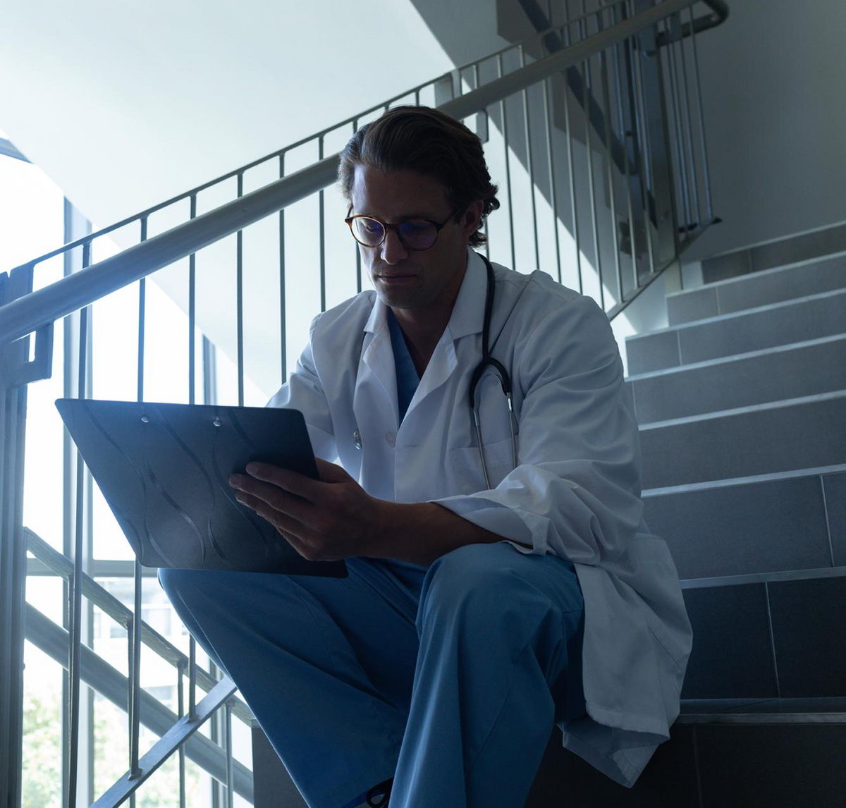 Nurse in navy scrubs resting with his eyes closed in a hospital stairwell